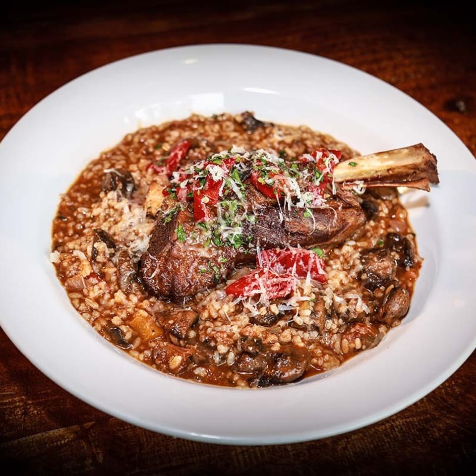 Braised lamb shank with risotto, mushrooms, red peppers, and grated cheese in a white bowl.
