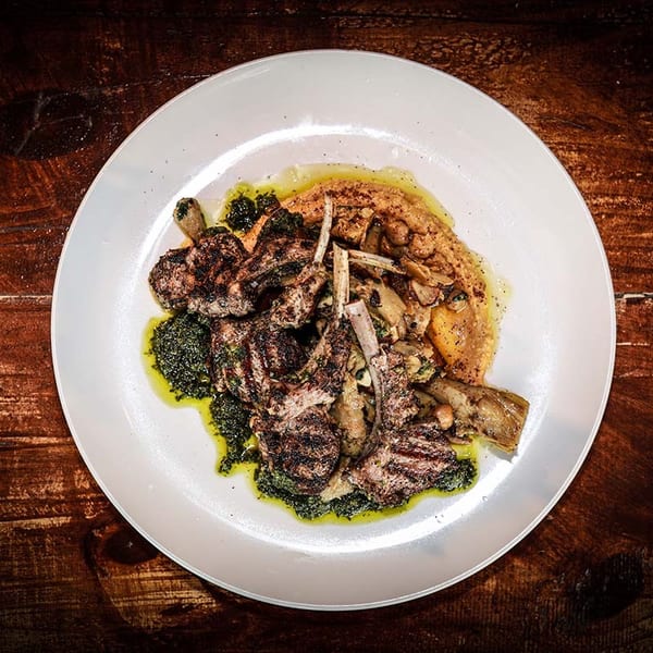 Grilled lamb chops with green sauce and a side of hummus and vegetables on a white plate.