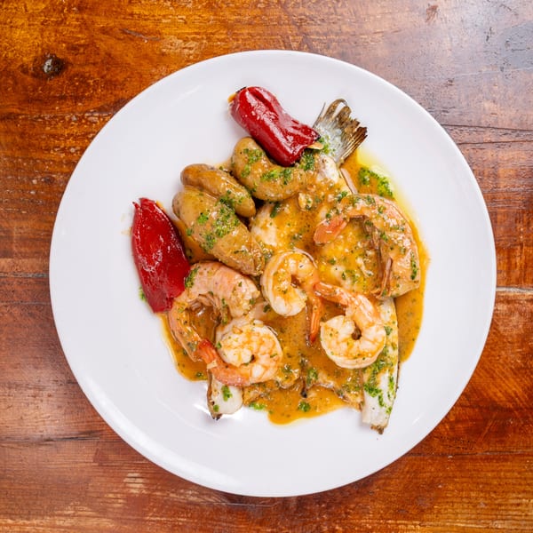 Plate of shrimp, roasted potatoes, red peppers, and fish drizzled with herb sauce on a rustic wooden table.
