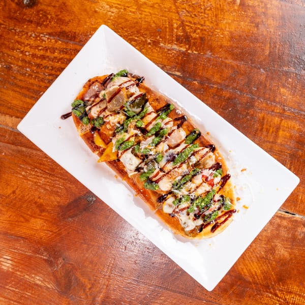 Loaded flatbread topped with chicken, vegetables, melted cheese, green pesto, and balsamic glaze on a white plate.