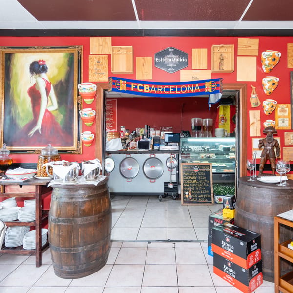 Colorful Spanish restaurant interior with a service counter, wine barrels, Barcelona scarf, paintings, ceramics, and Estrella Galicia beer boxes.
