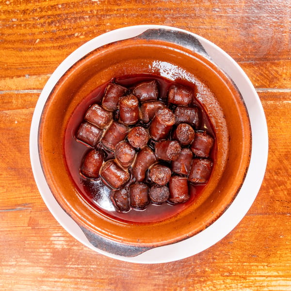 Overhead view of sliced sausage pieces simmering in dark sauce inside a rustic brown ceramic dish on a wooden table.