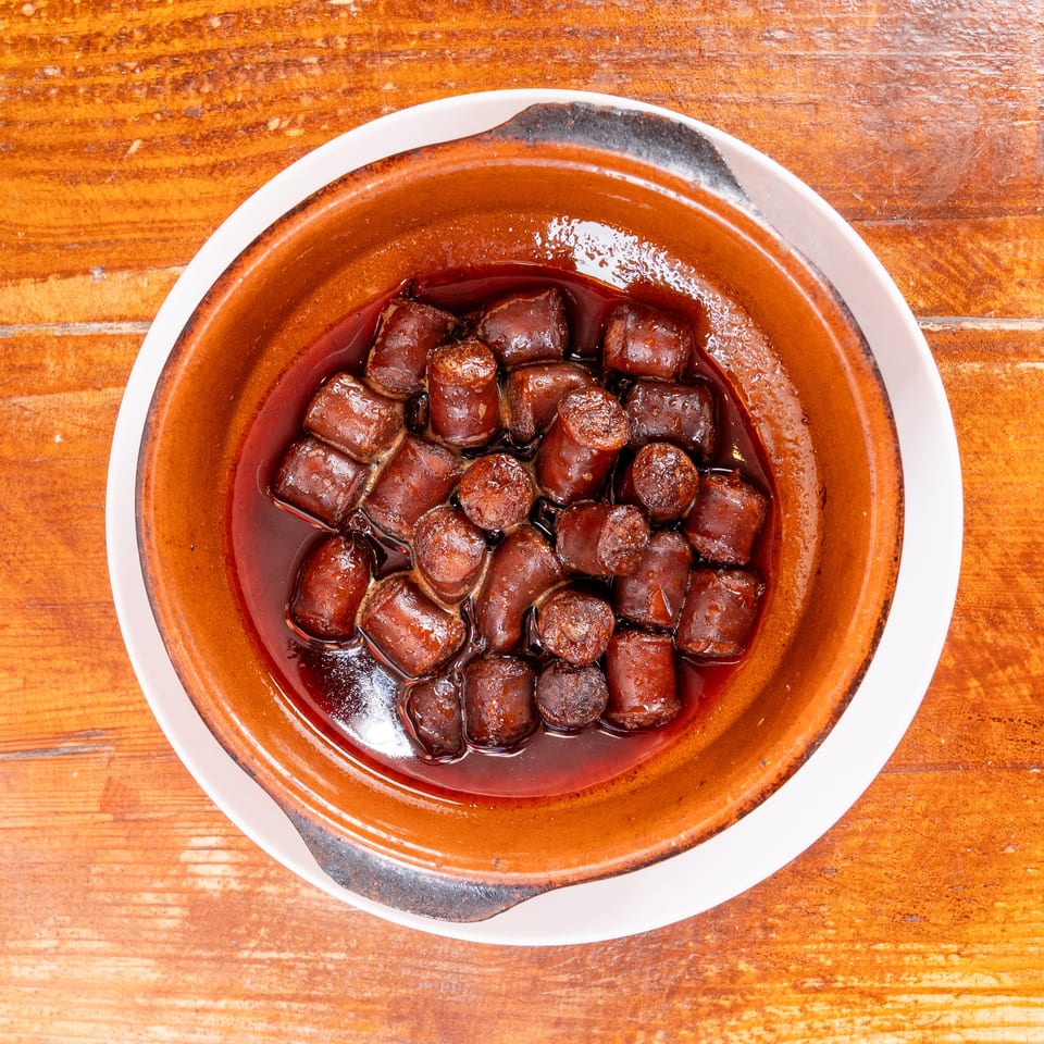Overhead view of sliced sausage pieces simmering in dark sauce inside a rustic brown ceramic dish on a wooden table.