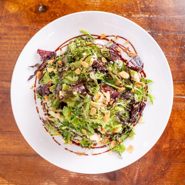 Mixed green salad with beet pieces, crumbled cheese, sliced almonds, and balsamic drizzle on a white plate.