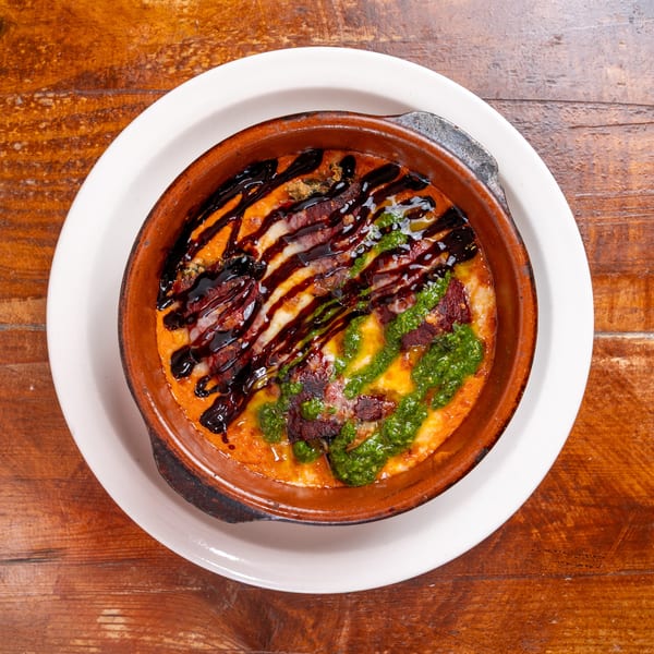 Overhead view of a colorful baked dish in a terracotta bowl, topped with sliced meat, green herb sauce, and dark glaze.