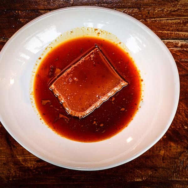 A square of flan dessert in a pool of caramel sauce on a white plate.