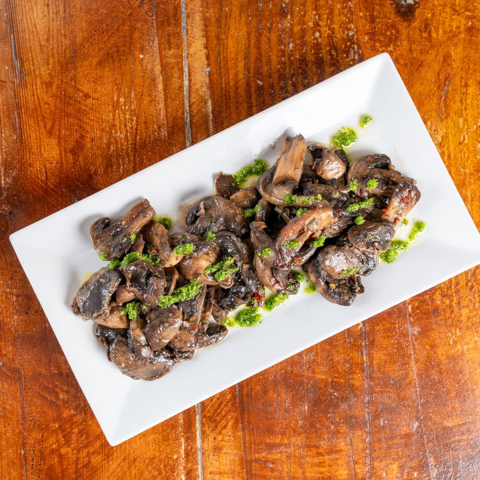 Sautéed mushrooms drizzled with green herb sauce on a white rectangular plate atop a rustic wooden table.