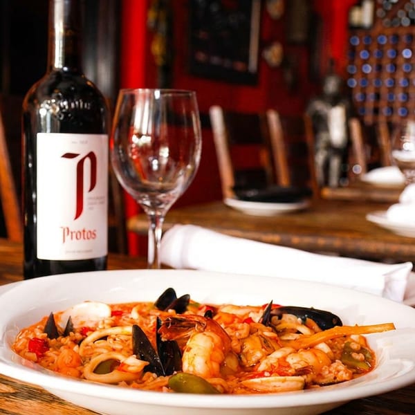 Seafood paella, a bottle of red wine, and wine glasses on a rustic wooden table.