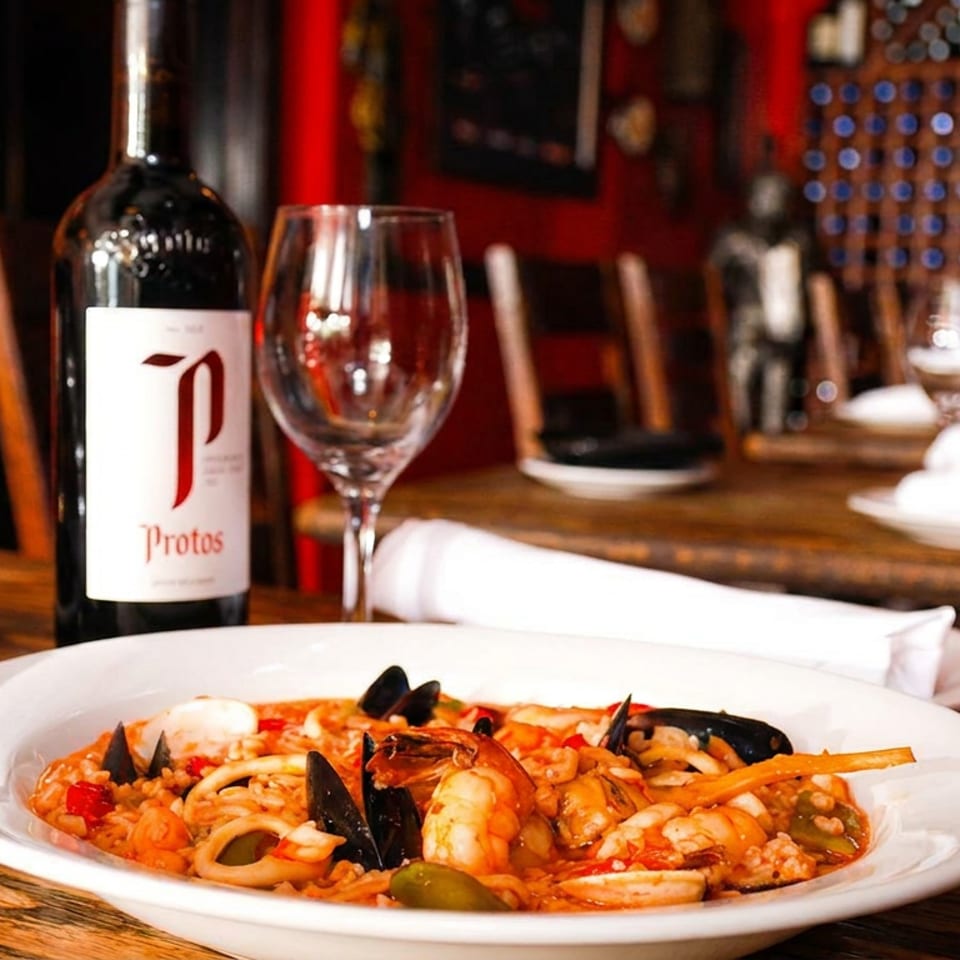 Seafood paella, a bottle of red wine, and wine glasses on a rustic wooden table.