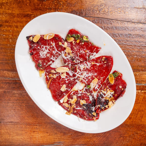 Thinly sliced beef carpaccio topped with shaved cheese, toasted almonds, herbs, and sauce on a white plate.