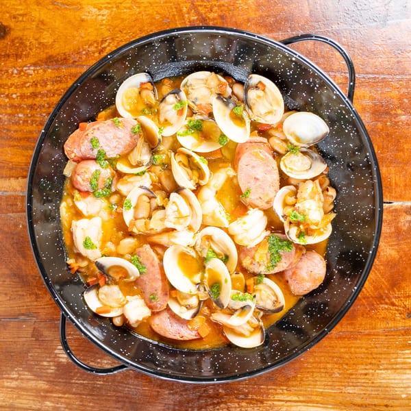 Overhead view of clams, sausage, shrimp, and vegetables in a savory broth, served in a black skillet on a wooden table.