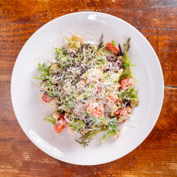 Fresh mixed green salad with tomatoes, olives, chickpeas, and shredded cheese served on a white plate atop a wooden table.