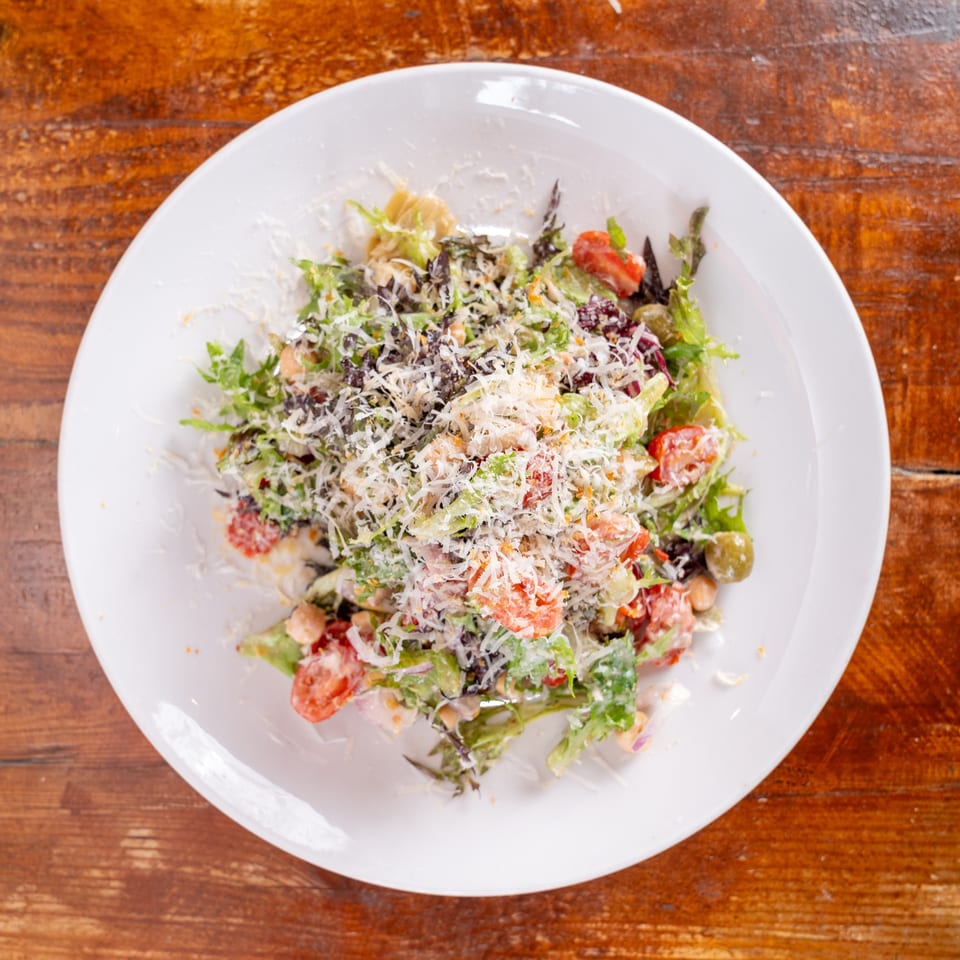 Fresh mixed green salad with tomatoes, olives, chickpeas, and shredded cheese served on a white plate atop a wooden table.