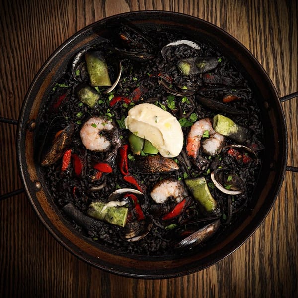 Black paella with shrimp, mussels, clams, red peppers, and a dollop of aioli in a pan.