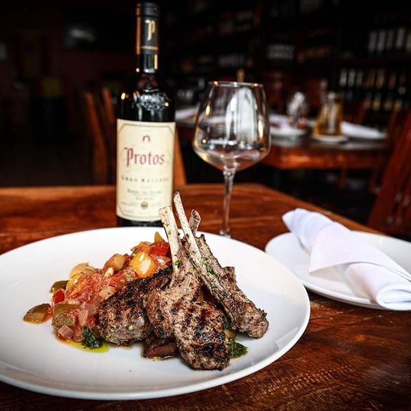 Grilled lamb chops with vegetables, a bottle of Protos wine, and an empty wine glass on a wooden table.