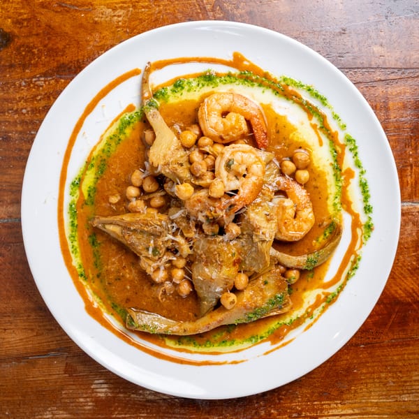 Plate of shrimp, fish, and chickpeas in a rich orange sauce, garnished with green herb oil on a wooden table.