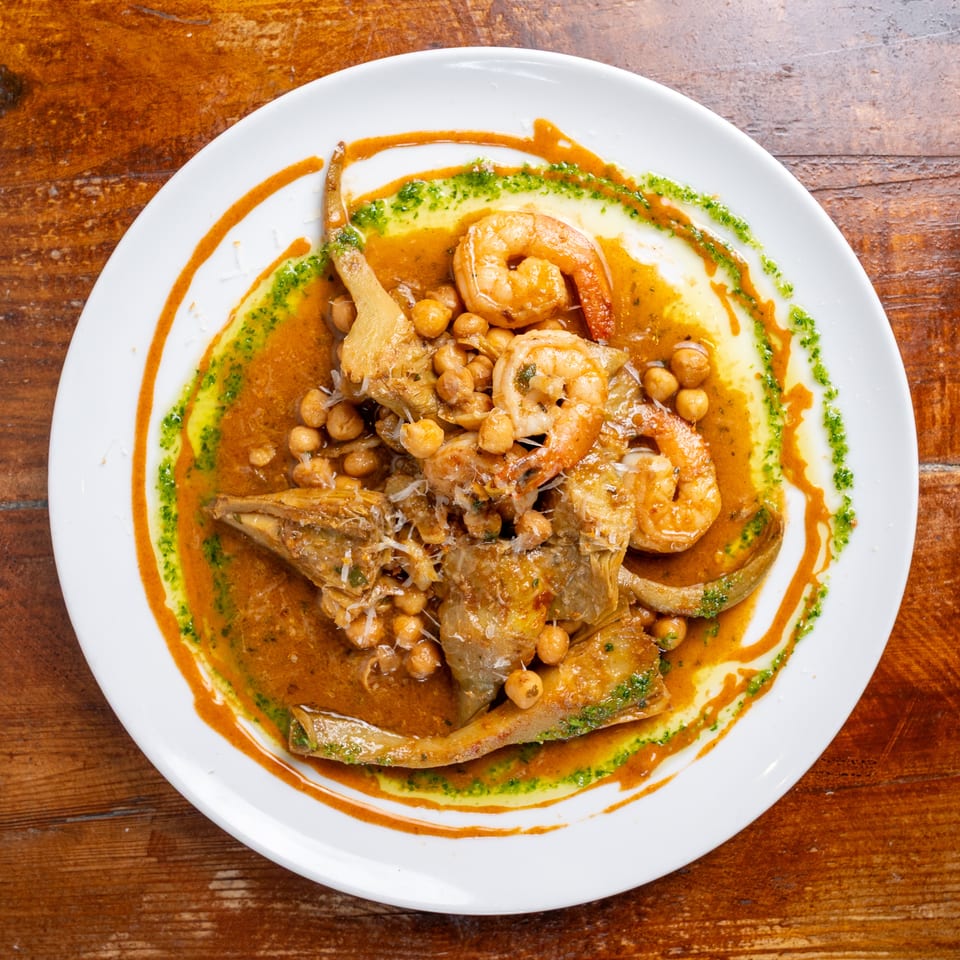 Plate of shrimp, fish, and chickpeas in a rich orange sauce, garnished with green herb oil on a wooden table.
