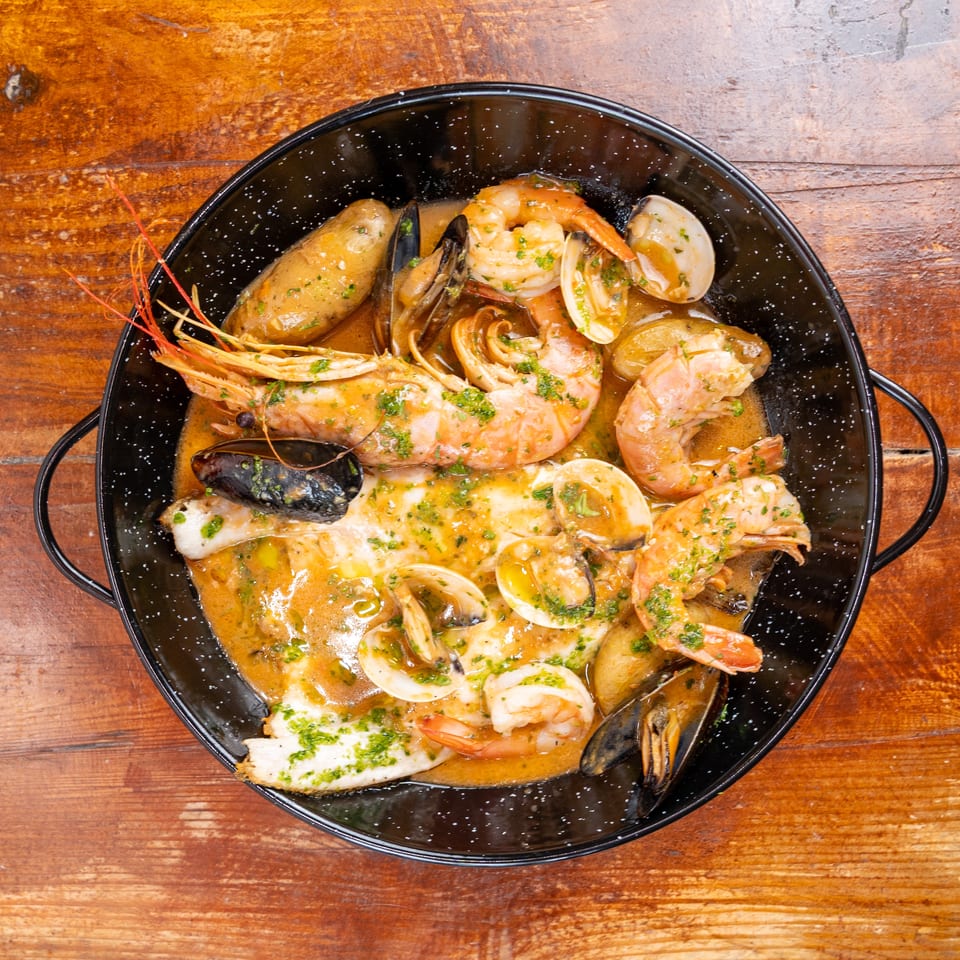 Overhead view of shrimp, mussels, clams, and fish in a herb-butter sauce served in a black pan on a wooden table.