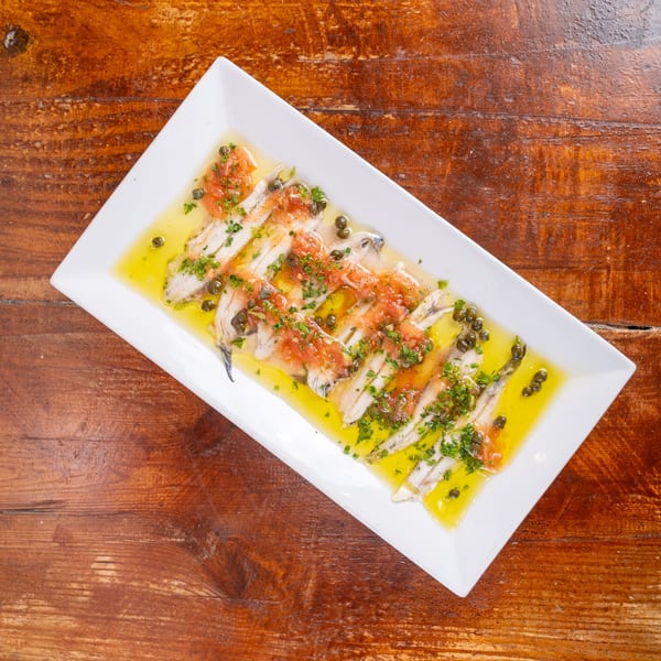 Sliced fish topped with tomatoes, capers, herbs, and olive oil on a white platter over a rustic wooden table.