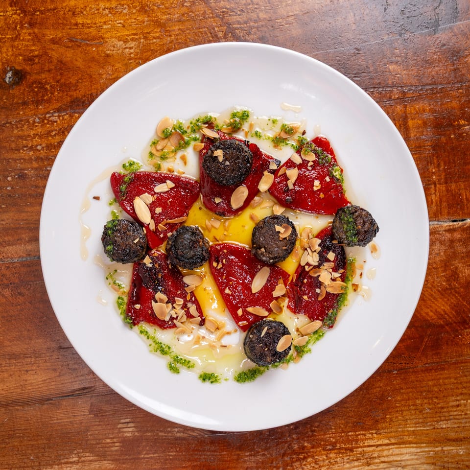 Plated roasted red peppers and black olives topped with sliced almonds, herbs, and olive oil on a white dish.