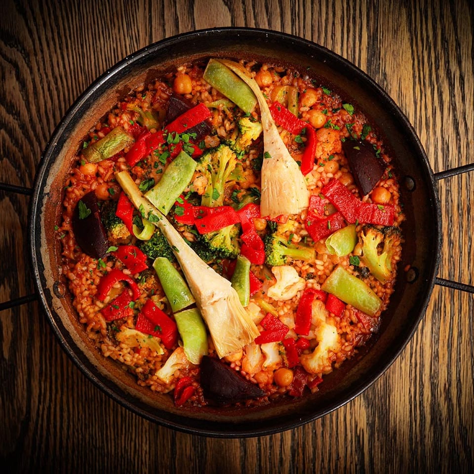 Vegetable paella with red peppers, broccoli, and artichoke hearts in a black pan on a wooden table.