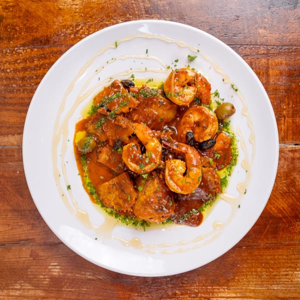 Plated shrimp and vegetables in a rich tomato sauce, garnished with herbs, olives, and drizzled oil on a wooden table.