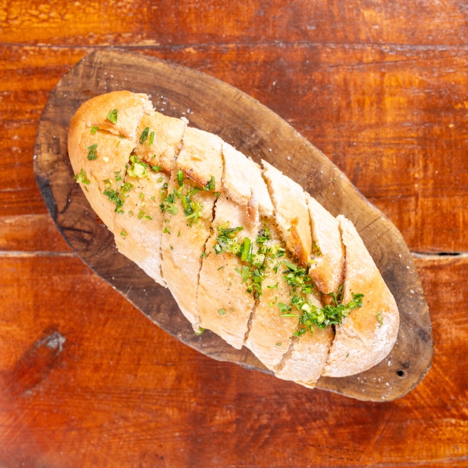 Sliced garlic bread topped with chopped herbs, served on a rustic wooden board atop a wooden table.