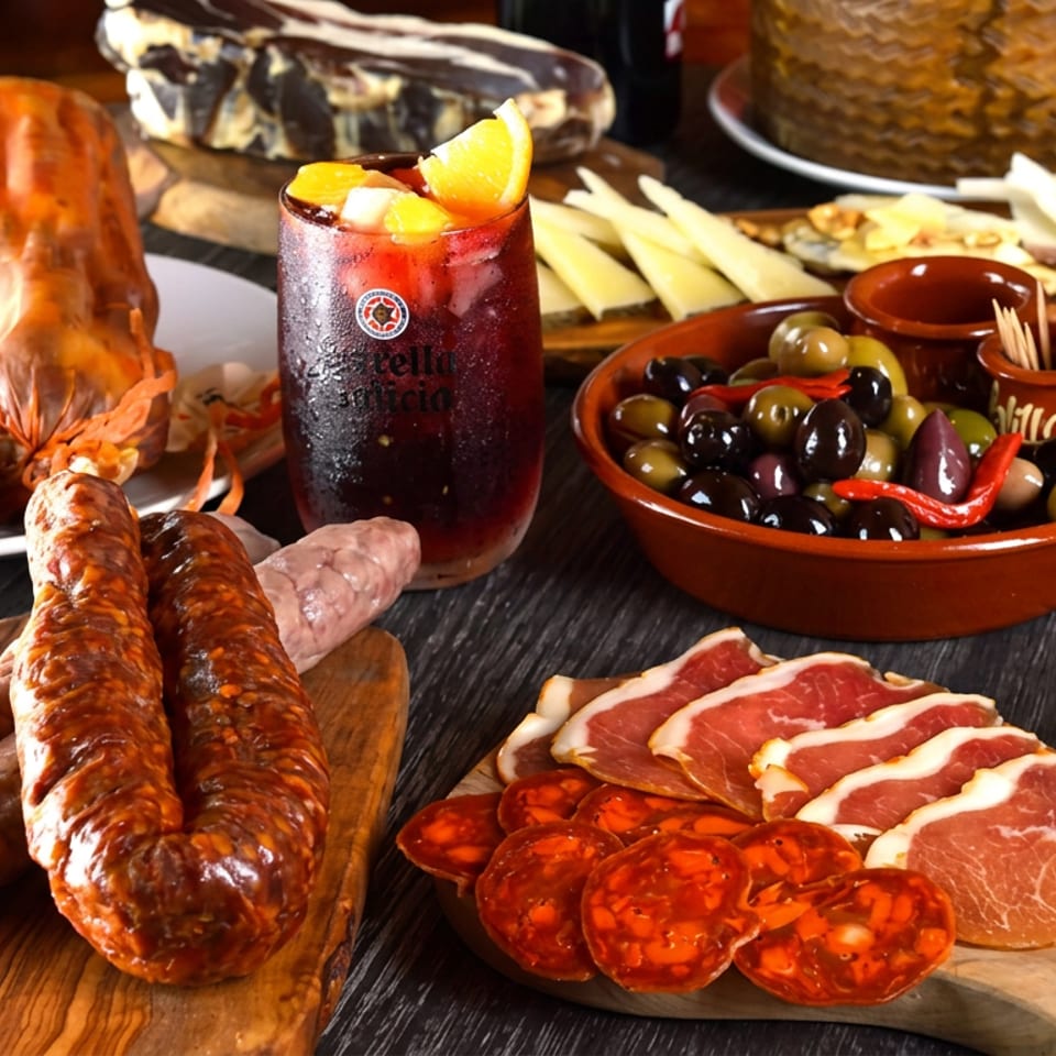 Assortment of Spanish tapas, including various sausages, cured meats, cheeses, olives, and a drink.