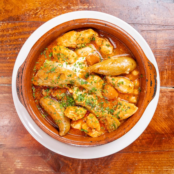 Clay dish of chicken and vegetables in rich orange sauce, garnished with herbs, on a wooden table.