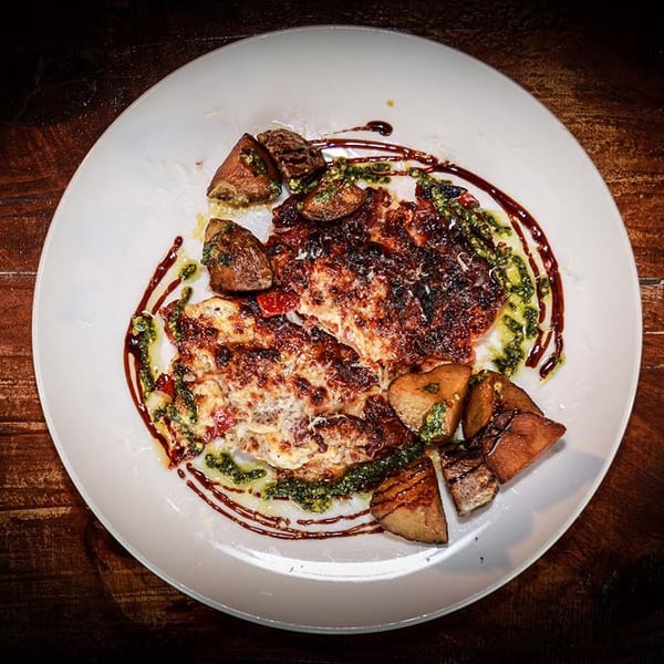 A white plate holds two cheesy baked chicken breasts with roasted potatoes, drizzled with pesto and balsamic glaze.