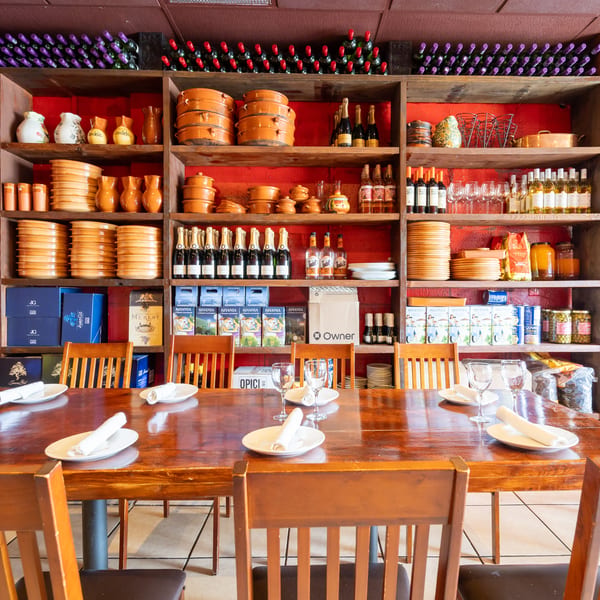 Restaurant dining room with set tables, wooden chairs, wine bottles, terracotta dishware, and colorful shelves against a red wall.