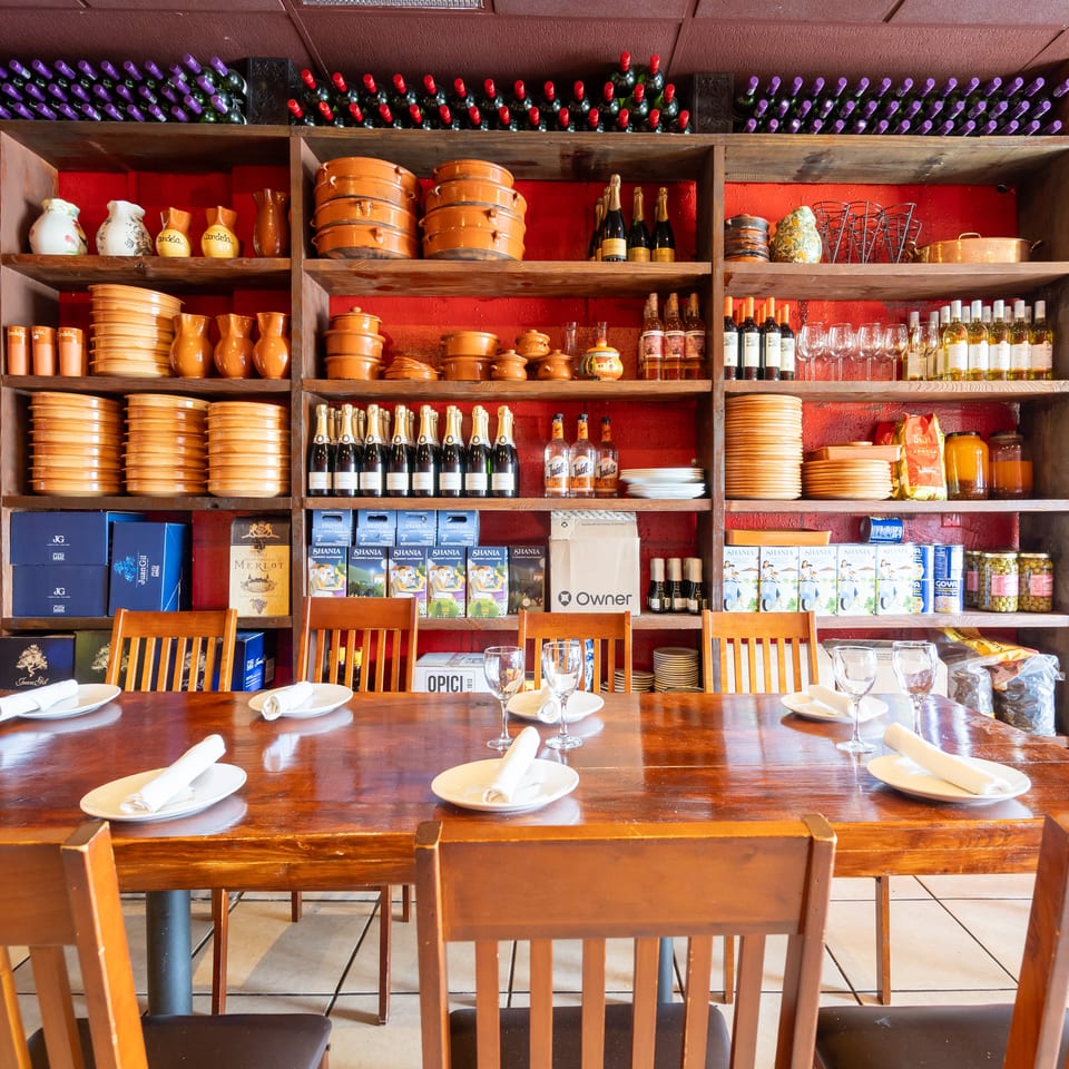Restaurant dining room with set tables, wooden chairs, wine bottles, terracotta dishware, and colorful shelves against a red wall.