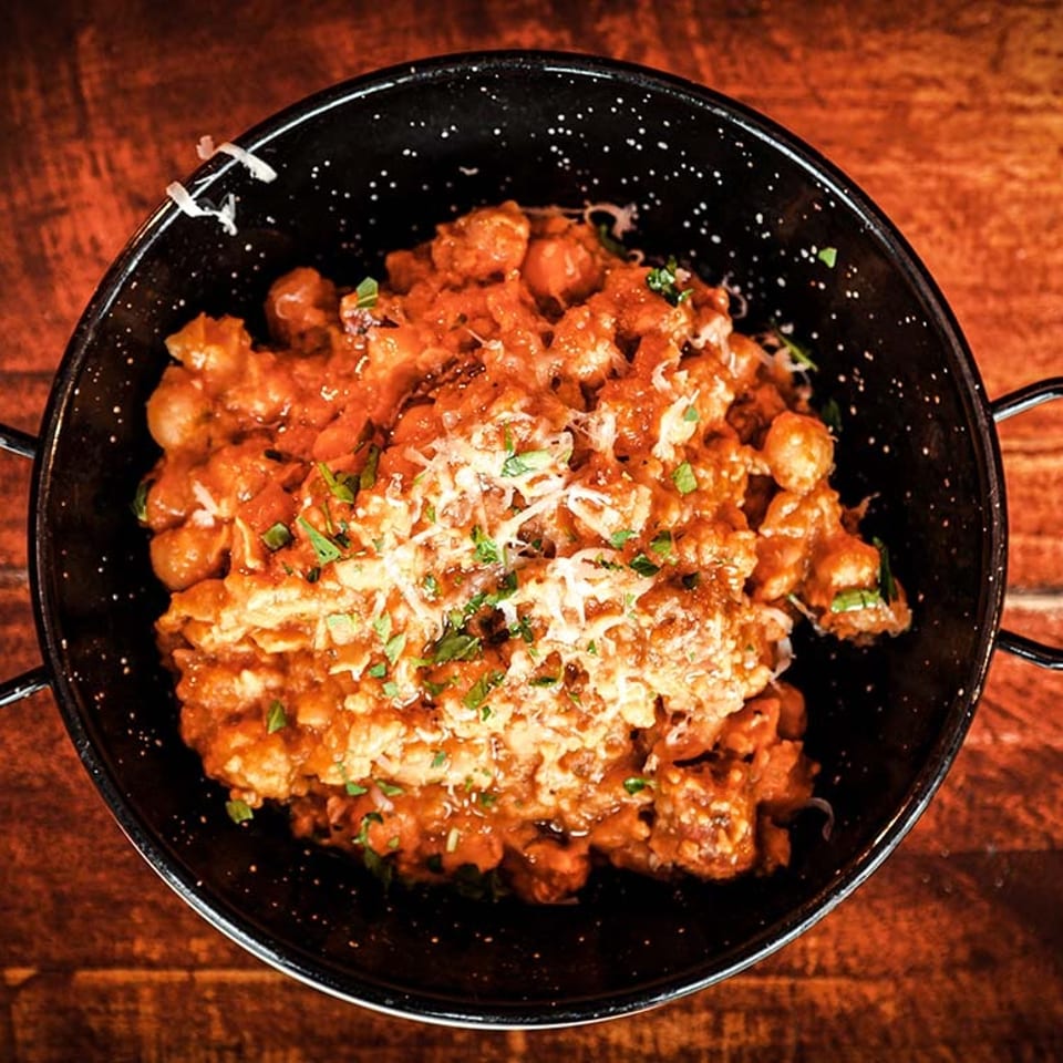 Hearty stew with chickpeas, meat, and rice, topped with cheese and herbs in a black pan.