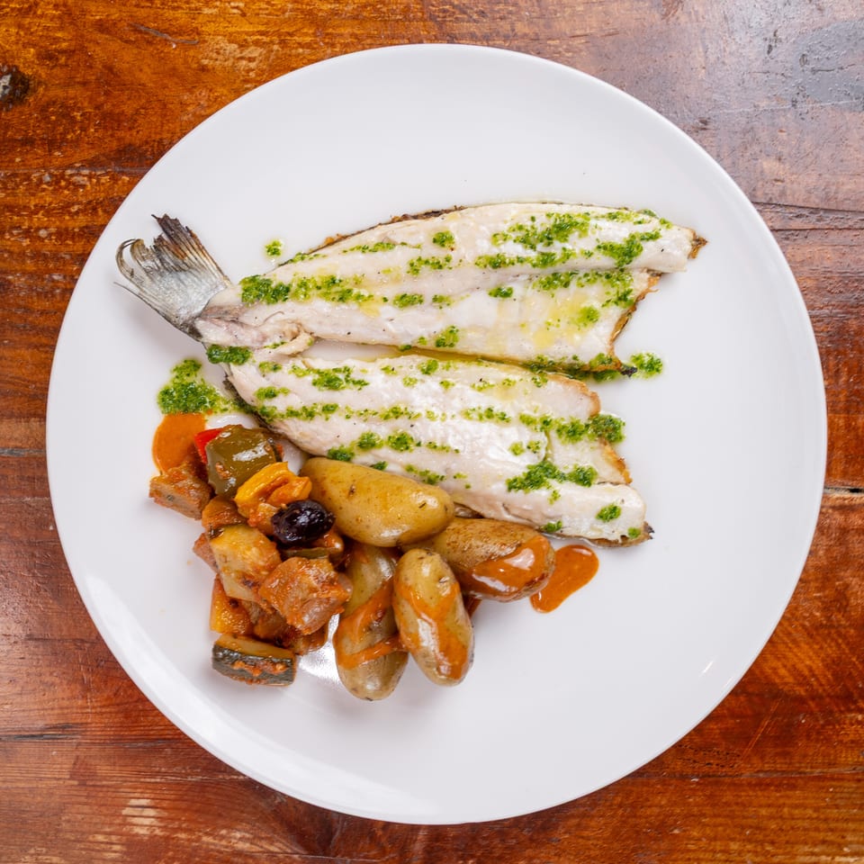 Grilled fish fillets topped with green herb sauce, served with roasted potatoes and vegetables on a white plate.